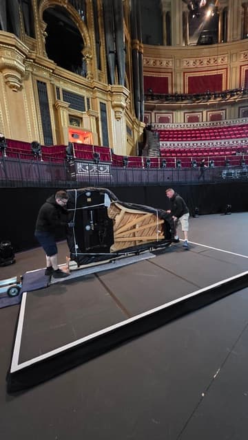 JJS Pianos crew rolling a wrapped grand piano on stage at a concert hall